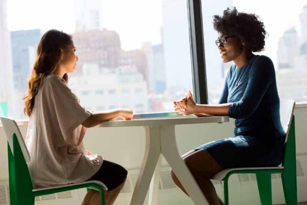 Two women talking at a white table in front of a window